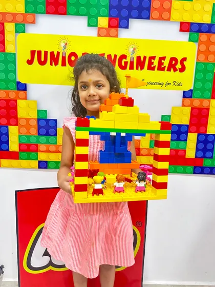 A young girl in a pink dress proudly holds up the colorful LEGO pandal she constructed.