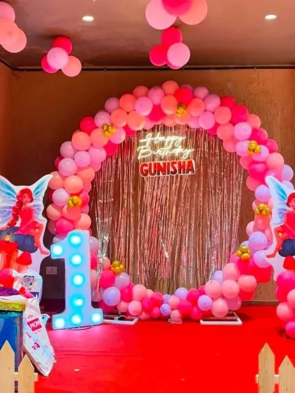 A fairy-themed first birthday party for Gunisha. The centerpiece is a large circular balloon arch in shades of pink, set against a shimmering backdrop, with fairy cutouts and a light-up number '1'.