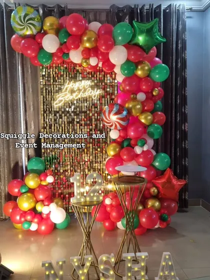 A festive birthday decoration featuring a gold shimmer wall, a 'Happy Birthday' neon sign, and a colorful red, green, and gold balloon arch.