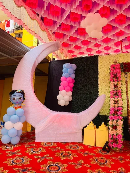 A baby shower stage featuring a moon prop with a Krishna cutout, set under a pink and white pandal ceiling.
