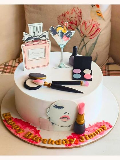 A chic makeup-themed cake featuring edible lipstick, eyeshadow, brushes, and a Miss Dior perfume bottle.