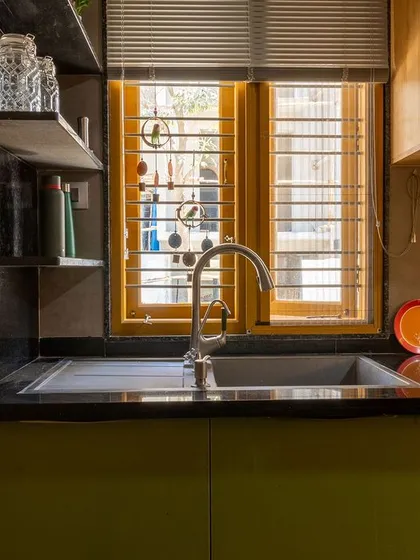 The kitchen combines functionality with natural aesthetics. Olive green laminate cabinets are set against exposed brick walls and a black granite countertop, showing how modern needs can be met within a sustainable framework.