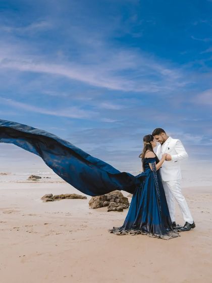 The navy blue infinity gown on a beautiful sandy beach. The deep blue of the dress contrasts stunningly with the groom's white suit and the bright sky.
