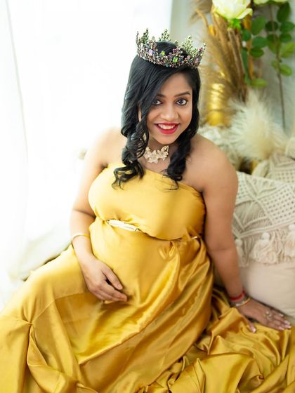 A bright and cheerful portrait of a mom-to-be in a golden satin dress and a crown, smiling happily in our cozy studio setup.