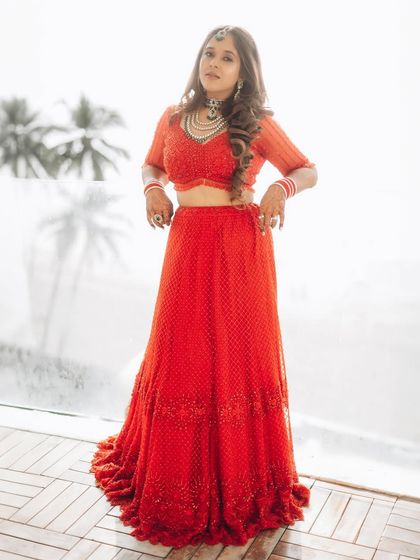 A full-length bridal portrait against a bright window. The backlighting creates a soft, ethereal glow, highlighting her silhouette and the flow of her lehenga.