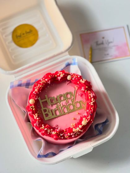 A vibrant pink birthday bento with a decorative red border and a modern gold "Happy Birthday" acrylic topper.