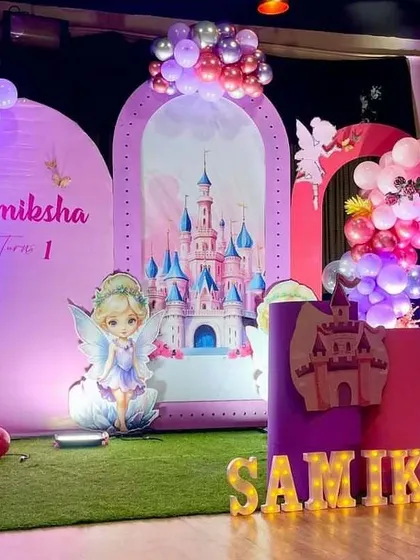 A magical fairy and castle themed birthday stage. This decoration uses arched backdrops with castle imagery, character cutouts of fairies, and balloon arrangements in pink and purple hues to create a fairytale setting.