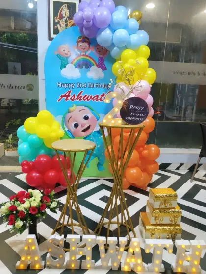 A bright and colorful Cocomelon theme for a 2nd birthday party. This setup features a custom character backdrop, a vibrant multi-colored balloon garland, and light-up letters spelling the birthday boy's name, 'ASHWATH'.