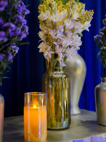 A simple yet elegant detail of white tuberoses in a golden vase, with a candle casting a warm glow against the royal blue backdrop.