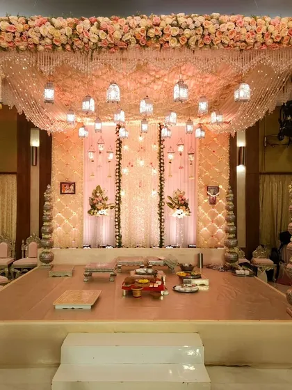 This is a beautiful pastel wedding mandap, featuring a canopy of draped fabric and hanging lanterns. The structure is supported by ornate silver pillars, and the entire stage is decorated with a thick border of pink and white roses for a classic, romantic feel.