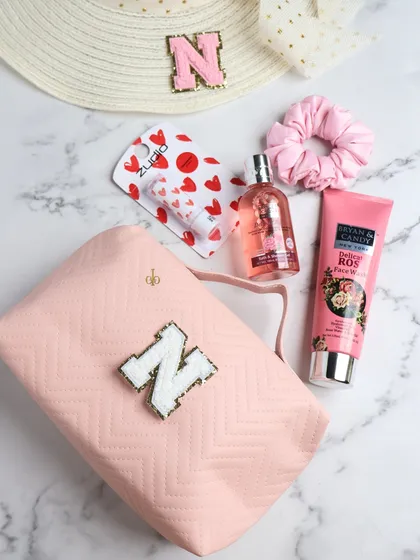 A flat lay showing our personalized pink pouch, perfect for carrying daily essentials like perfume, face wash, and a scrunchie. It's paired with a matching initialed sun hat.
