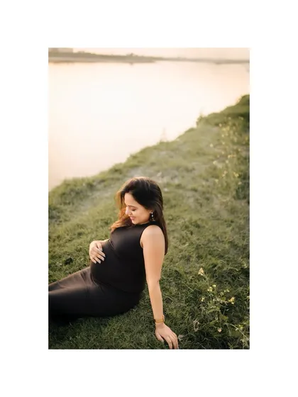 A wider perspective of the serene lakeside scene, showing the expecting mother enjoying a peaceful moment in nature. The calm water and soft light create a tranquil atmosphere.