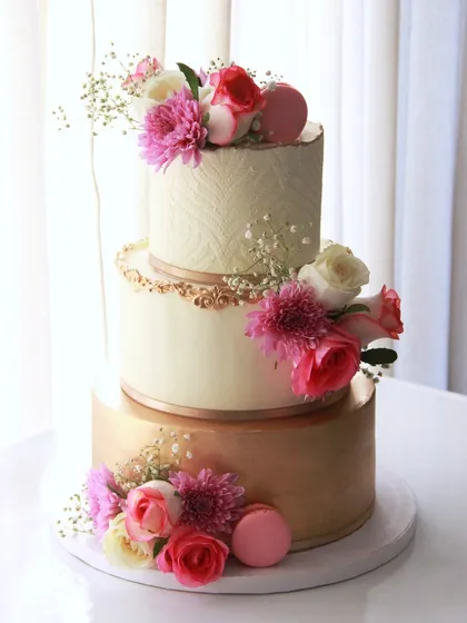 An elegant and minimalist three-tiered engagement cake. The design combines a brushed gold bottom tier, a smooth middle tier, and a stenciled top tier, all tied together with fresh flowers and macarons.