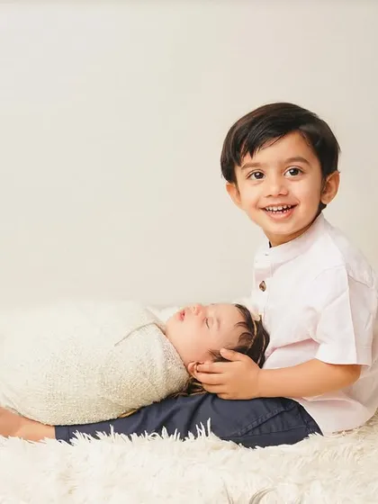A proud big brother smiles for the camera while his new sibling sleeps on his lap. He's already a natural at his new role.