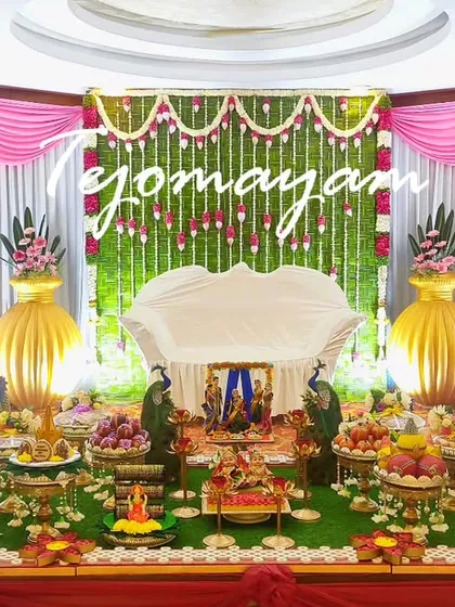 A baby shower setup with a backdrop of hanging green garlands and pink drapes. The two large golden pots on either side add a touch of grandeur.