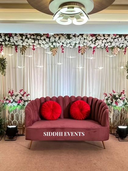 A sophisticated stage design with a dark pink scalloped sofa, a canopy of white flowers with gold leaf accents, and hanging bird decorations for a whimsical touch.
