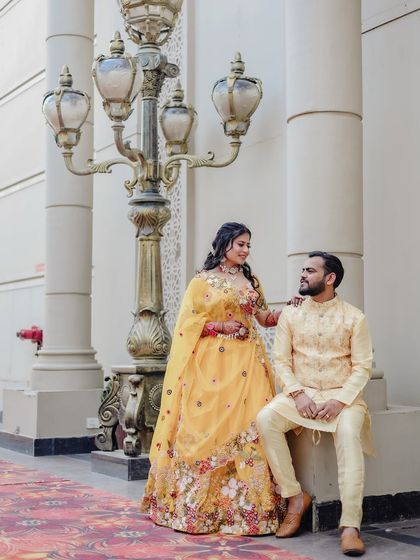 A full-length couple portrait by an ornate lamp post, showcasing their coordinated yellow outfits for the haldi function.