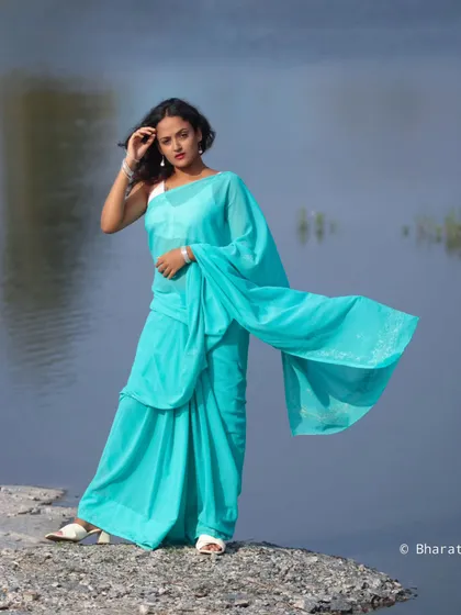 A striking outdoor shot of a model in a flowing turquoise saree against a calm water background. The wind catching the fabric creates a sense of drama and movement.