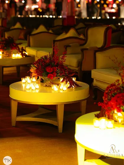 A cozy lounge corner with elegant seating, where the tables are lit by the warm glow of candles surrounding a rich, red floral arrangement.