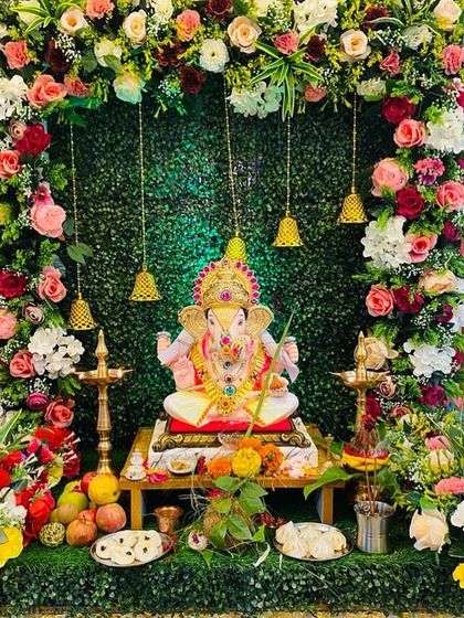 A beautiful Ganpati setup with a backdrop of artificial grass, framed by a dense arch of colorful flowers. The hanging temple bells add a traditional and auspicious element to the decor.