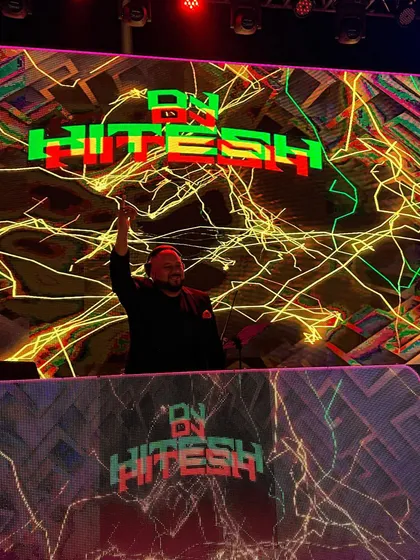 One hand up, feeling the energy from the crowd. The pixelated, multi-color visuals on the screen add to the dynamic, high-tech atmosphere of the party.
