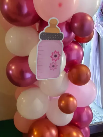 A detail of a balloon pillar for a baby girl's welcome, featuring a cute milk bottle cutout amidst pink, white, and chrome balloons.