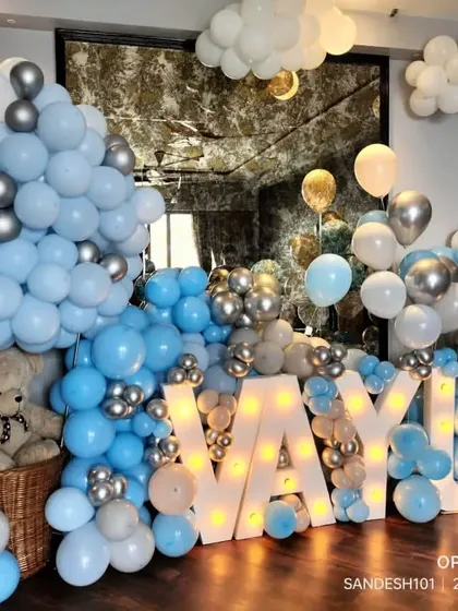 A cute and cozy baby welcome decoration. The setup features large marquee letters spelling "VAYU," surrounded by blue, white, and silver balloons and teddy bears.