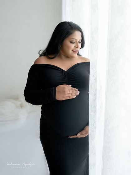 A simple and elegant solo portrait. The mom-to-be in a classic black gown stands by the window, the soft light beautifully sculpting her silhouette.