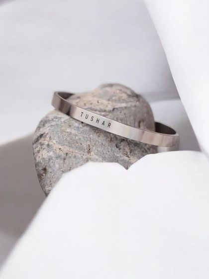 A minimalist shot of a silver cuff with the name 'TUSHAR', resting on a stone against a clean white background.