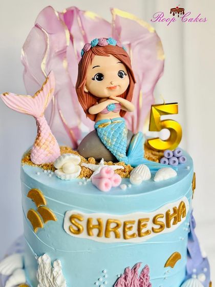 A detailed close-up of the adorable, handcrafted mermaid topper. Her tail shimmers, and she sits among edible sand and seashells, ready for a magical underwater party.