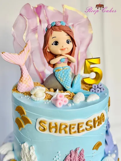 A detailed close-up of the adorable, handcrafted mermaid topper. Her tail shimmers, and she sits among edible sand and seashells, ready for a magical underwater party.