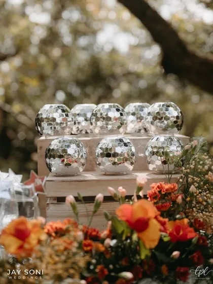 Small disco ball candle holders add a touch of sparkle to a rustic wooden crate, set against a backdrop of vibrant orange flowers.