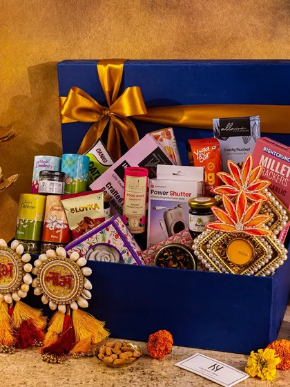 This royal blue hamper box is overflowing with goodies, styled with traditional 'Shubh Labh' decor. The warm, moody lighting creates a cozy and inviting atmosphere.