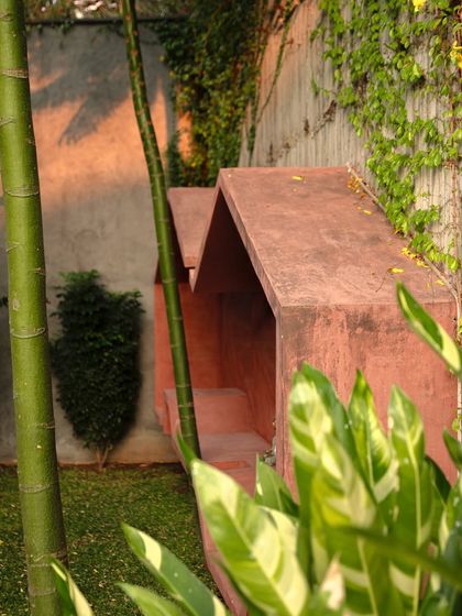 A sculptural seating nook with a geometric, angular form, finished in a warm red oxide. The piece is nestled among bamboo and other tropical plants, creating a private and artistic corner in the garden.