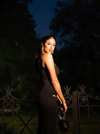 A profile shot taken at dusk, using flash to create a dramatic effect. The lighting highlights the model's silhouette and the sleek design of the black dress against the dark background.