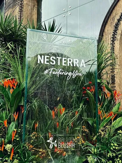 A clear acrylic welcome sign for the Nesterra event, placed amidst tropical plants and flowers. The transparent design creates a modern look while blending with the natural decor.