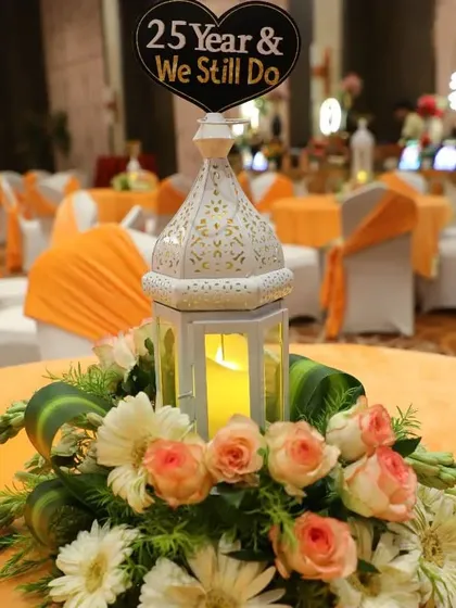 I create custom table centerpieces to match the event theme. This one was for a 25th anniversary party, featuring a white lantern with a candle, surrounded by fresh roses and daisies on an orange tablecloth.