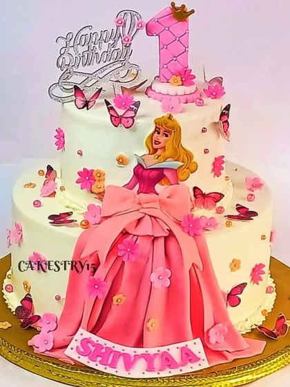 A regal two-tier Princess Aurora cake for a first birthday. The cake is decorated with a fondant princess figure, a flowing pink gown, and delicate butterflies.