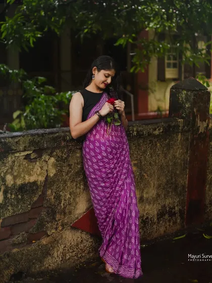 A full-length portrait in a rustic setting. The model leans against an old wall, creating a relaxed and natural pose that beautifully showcases the drape of her purple printed saree.