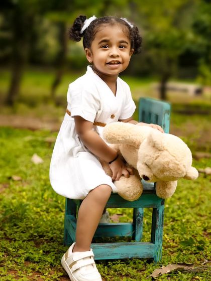 A solo portrait of a little girl sitting on a rustic blue chair, hugging her teddy bear in a park setting.