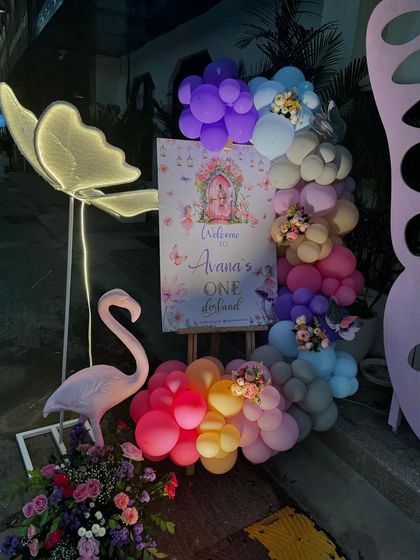 A welcome sign for "Avana's One-derland" decorated with a colorful balloon garland and flanked by a flamingo and an illuminated butterfly prop.