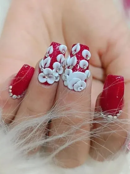 A close-up of our 3D floral nail art. Here, we've sculpted delicate white flowers on a bold red base, surrounded by tiny rhinestones for extra sparkle.