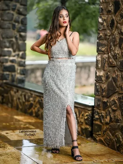 A high-fashion outdoor portrait featuring a unique silver tinsel two-piece set, creating a glamorous and textured look against a stone wall.