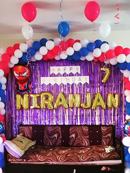 A classic Spiderman birthday party setup, perfect for a superhero fan's seventh birthday. The decoration features a red, white, and blue balloon arch over a couch, set against a purple shimmer curtain. A Spiderman foil balloon and the name "NIRANJAN" in gold letters make it clear who the hero of the day is.