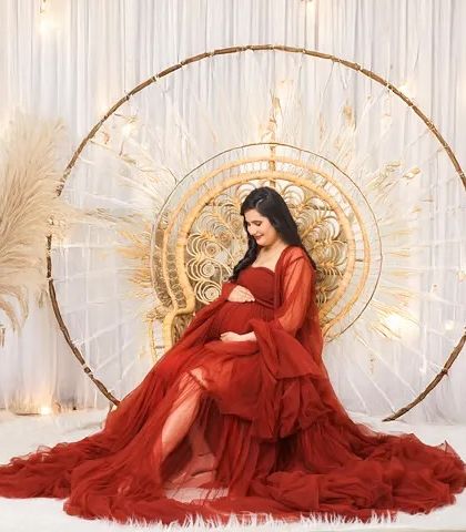 A radiant mom-to-be in a flowing red gown, seated on a peacock chair in a brightly lit studio. The boho-inspired setup with string lights adds a touch of warmth and magic.