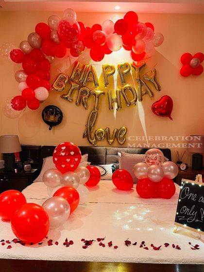 A classic red and silver themed birthday room decoration for a loved one. The setup includes 'Happy Birthday Love' foil balloons, an organic balloon arch over the headboard, and smaller balloon bouquets on the bed.
