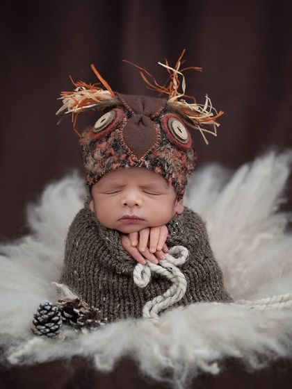 A wise little owl. This baby wears a creative, textured owl hat and is wrapped in a brown swaddle, sleeping in a soft white nest.