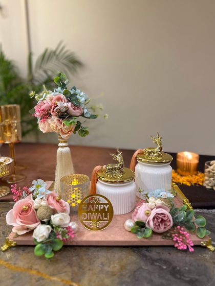 This Diwali hamper is designed to be a beautiful decor piece. It features a pink marble tray with two white ceramic jars topped with golden deer, a floral bouquet, and a 'Happy Diwali' tag, creating a complete and elegant gift set.