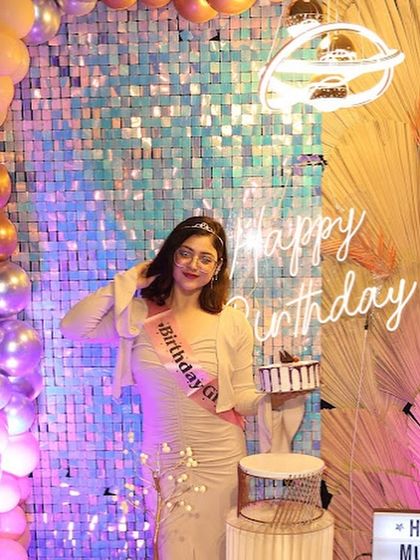 The birthday girl, wearing a "Birthday Girl" sash, poses with her cake. The iridescent sequin wall and soft lighting create a dreamy, celebratory atmosphere.