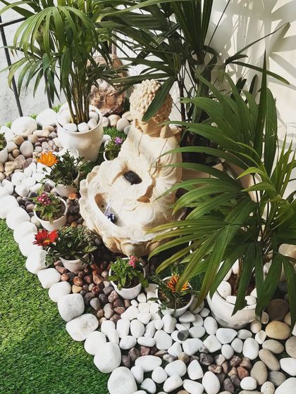 A serene corner of a garden featuring a Buddha water fountain surrounded by pebbles and small flowering plants.
